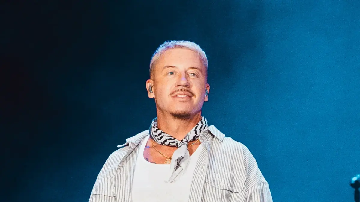Macklemore sur scène en concert, cheveux décolorés, foulard noir et blanc et chemise rayée, sur fond bleu (2026)