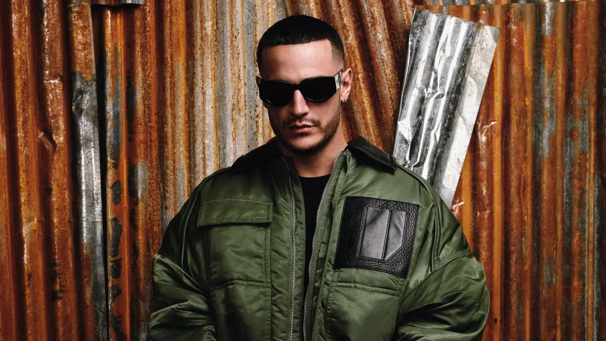 DJ Snake en blouson kaki et lunettes noires devant un mur de tôle rouillée, photo de 2025