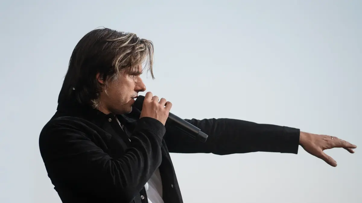 Orelsan chantant au micro sur scène, de profil, bras tendu, en veste noire, lors d'un concert en 2023