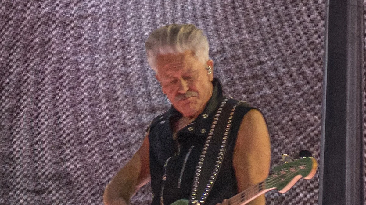 Un musicien de U2 aux cheveux gris, en gilet clouté noir, joue de la basse sur scène lors d'un concert en 2024