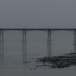 Visual extrated from the album "Without Sinking" by Hildur Gudnadottir : view of a bridge leading to an industrial island from the seashore, in a darken grey weather