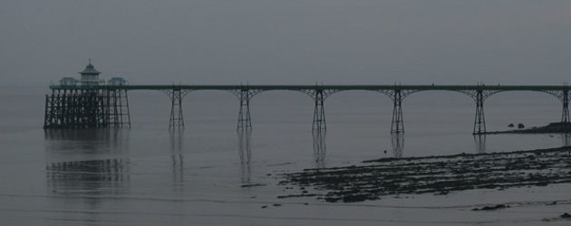 Visual extrated from the album "Without Sinking" by Hildur Gudnadottir : view of a bridge leading to an industrial island from the seashore, in a darken grey weather